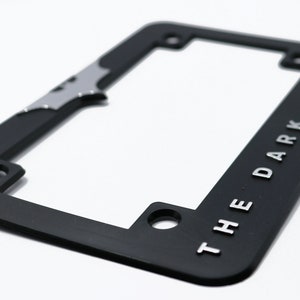 May include: Black license plate frame with the words "THE DARK KNIGHT" in silver lettering. The frame has a silver bat symbol and is designed to fit a standard U.S. license plate.