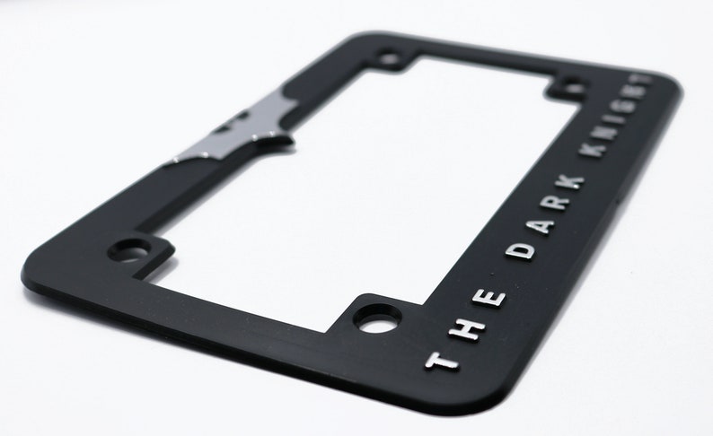 May include: Black license plate frame with the words "THE DARK KNIGHT" in silver lettering. The frame has a silver bat symbol and is designed to fit a standard U.S. license plate.
