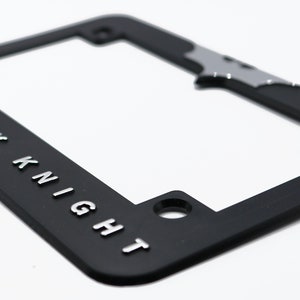 May include: Black license plate frame with the words "THE DARK KNIGHT" in silver lettering. A silver bat symbol is at the top. The frame has mounting holes and a rectangular shape.