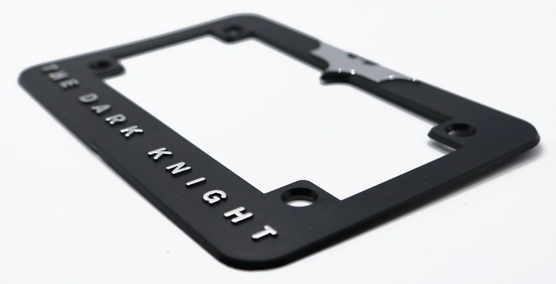 May include: Black license plate frame with the words "THE DARK KNIGHT" in silver lettering. A silver bat symbol is at the top. The frame has mounting holes and a rectangular shape.