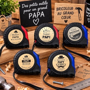 Peut inclure: Cinq mètres rubans noirs avec des inserts circulaires en bois personnalisés portant divers textes français et des icônes comme des chapeaux, des moustaches et des outils. Les mètres sont disposés sur une surface en bois avec une règle en métal.