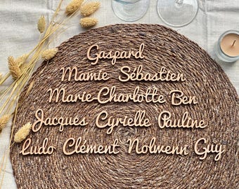 Peut inclure: Une collection de prénoms découpés au laser dans du bois avec une police cursive, incluant Gaspard, Mamie, Sébastien, Marie-Charlotte, Ben, Jacques, Cyrielle, Pauline, Ludo, Clément, Nolwenn et Guy, disposés sur un set de table en jute tressé.