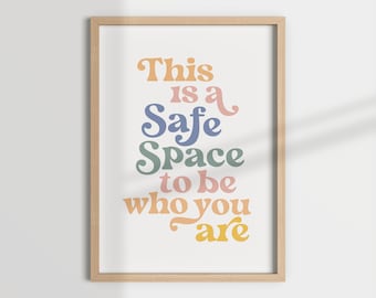 以下が含まれることがあります： 「This is a Safe Space to be who you are」というテキストが、カラフルなレトロなフォントで書かれた額装されたプリント。テキストはオレンジ、ピンク、ブルー、グリーンのシェードで書かれています。