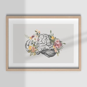 Puede incluir: Una impresión enmarcada de un cerebro humano con una corona floral de flores rosas, amarillas y blancas. El cerebro está representado en un estilo anatómico vintage.