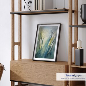 May include: A framed watercolor painting of a forest scene with trees and a path. The painting is in shades of blue, green, and yellow. The frame is black and the painting is on a wooden shelf.