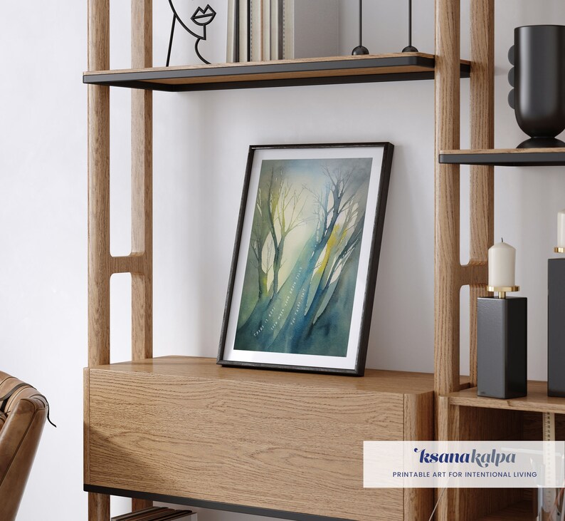 May include: A framed watercolor painting of a forest scene with trees and a path. The painting is in shades of blue, green, and yellow. The frame is black and the painting is on a wooden shelf.