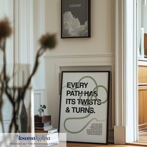 May include: Two framed prints hang on a white wall. The larger print has the text "EVERY PATH HAS ITS TWISTS & TURNS" in black and green. The smaller print features a grayscale image of clouds. The room has a wooden floor.