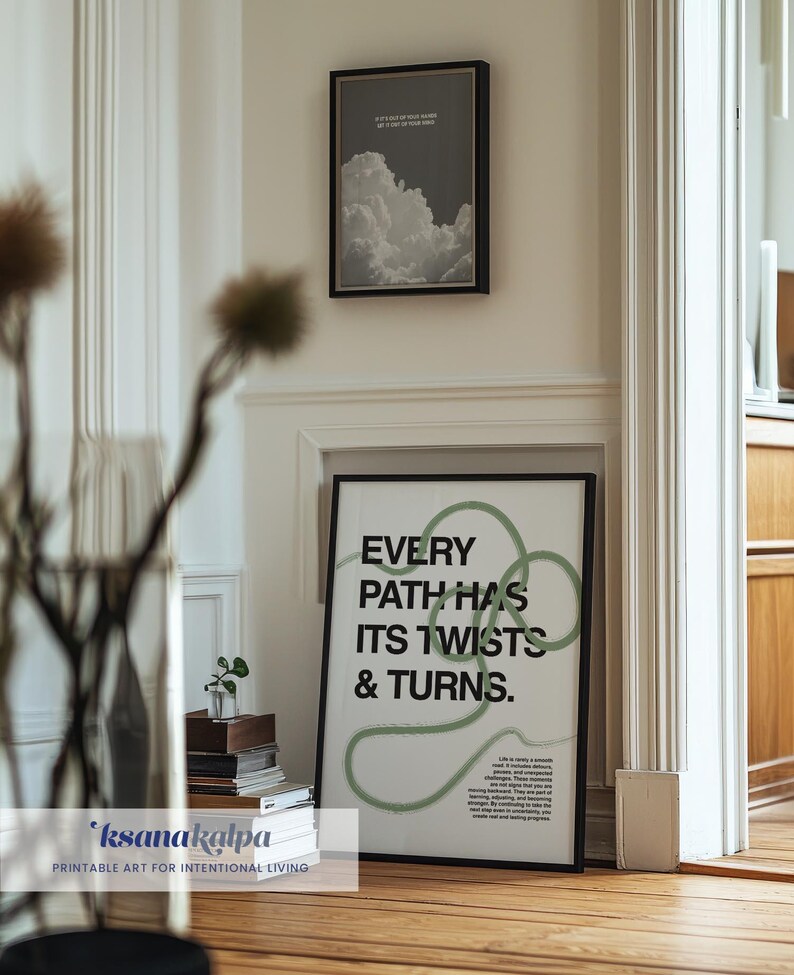 May include: Two framed prints hang on a white wall. The larger print has the text "EVERY PATH HAS ITS TWISTS & TURNS" in black and green. The smaller print features a grayscale image of clouds. The room has a wooden floor.