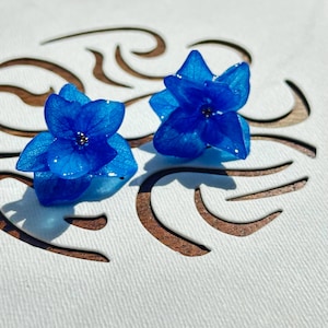 Puede incluir: Un par de pendientes azules en forma de flor. Los pendientes están hechos de resina y tienen un aspecto delicado y translúcido.