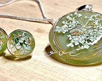 Puede incluir: Un conjunto de joyas de resina verde y blanca con una cadena de plata y detalles dorados. El colgante y los pendientes presentan delicadas flores blancas prensadas en la resina.