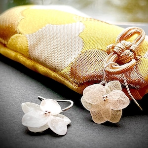 May include: Close-up of delicate, flower-shaped earrings with silver hooks. The earrings are a soft, translucent white with tiny bead accents. They rest on a dark surface near a yellow and gold patterned fabric.