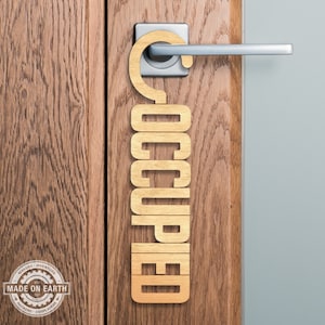 Puede incluir: Un colgador de puerta de madera con la palabra "OCUPADO" escrita en letras verticales. El colgador está sujeto a una puerta con un pomo de puerta plateado.