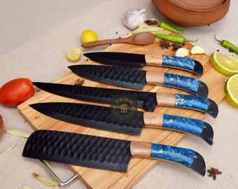 May include: Set of five black kitchen knives with blue and gold patterned handles on a wooden chopping board. The knives have a textured blade and are arranged in a row.
