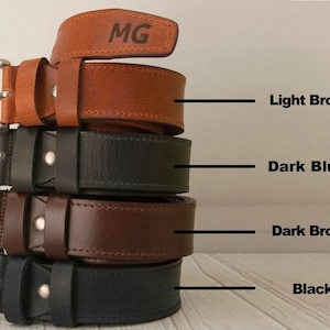 May include: A stack of four leather belts with silver-toned metal buckles, featuring colours labelled as Light Brown, Dark Blue, Dark Brown, and Black. The top light brown belt is engraved with the letters MG. Each belt has a single metal rivet.