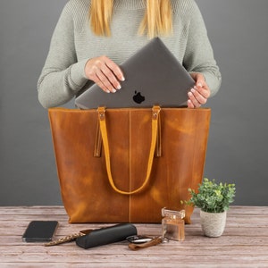 May include: A large tan leather tote bag with two shoulder straps and a vertical center seam. The bag is open at the top, revealing a dark gray laptop with a visible apple logo. The exterior features a smooth, matte finish with visible stitching.