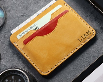 May include: A tan leather card holder with a stitched edge and the name "LIAM" embossed on the front. A red credit card and a white credit card are visible inside the holder.