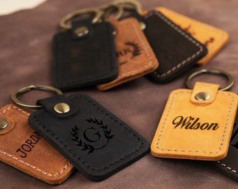 May include: A collection of six leather keychains with different engravings. The keychains are brown, black, and tan. The engravings include names, initials, and a wreath with a letter G.