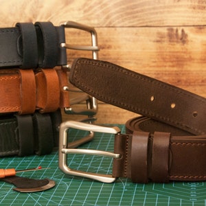 May include: A collection of leather belts in black, tan, and dark brown, each featuring a silver-toned metal buckle and two leather loops. A metal hole punch tool and a seam ripper are positioned near the belts on a green cutting mat.