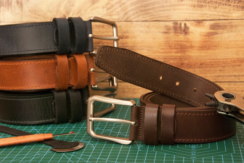May include: A collection of leather belts in black, tan, and dark brown, each featuring a silver-toned metal buckle and two leather loops. A metal hole punch tool and a seam ripper are positioned near the belts on a green cutting mat.