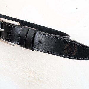 May include: A black leather belt featuring a silver-toned metal buckle and two matching leather loops. The belt strap has visible grey stitching along the edges and a debossed emblem on the pointed tip displaying the letter K inside a wreath.