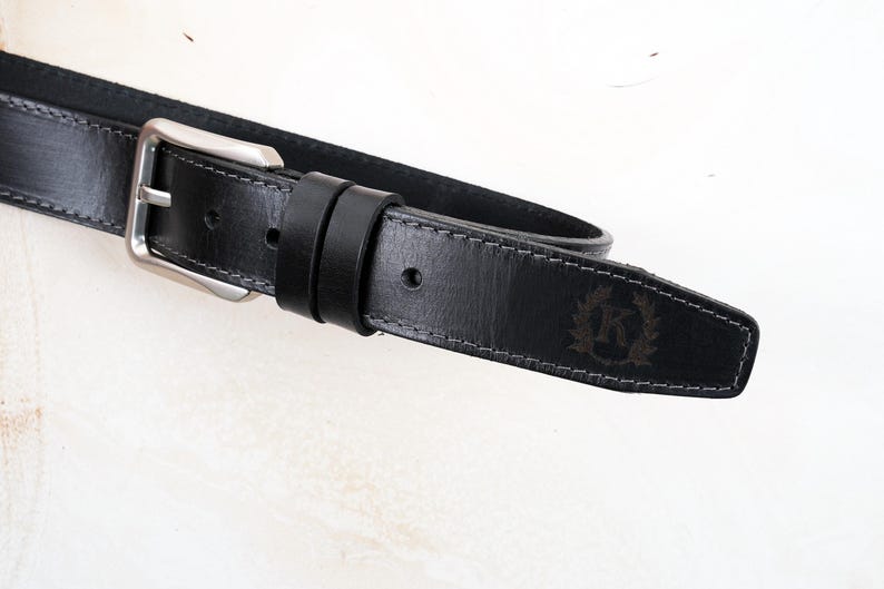 May include: A black leather belt featuring a silver-toned metal buckle and two matching leather loops. The belt strap has visible grey stitching along the edges and a debossed emblem on the pointed tip displaying the letter K inside a wreath.