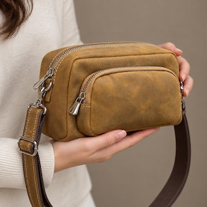 May include: A rectangular mustard-brown leather crossbody bag featuring a smooth matte finish, a front zippered pocket, and a main compartment with silver-toned metal zippers. It includes a wide, adjustable brown fabric shoulder strap.