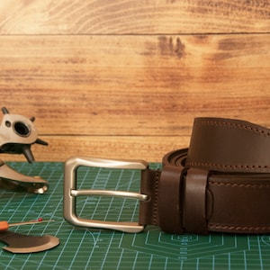 May include: A dark brown leather belt with a silver-toned metal buckle and matching stitching. Beside the belt lies a metal rotary leather hole punch tool and a small orange-handled seam ripper on a green cutting mat with grid markings.