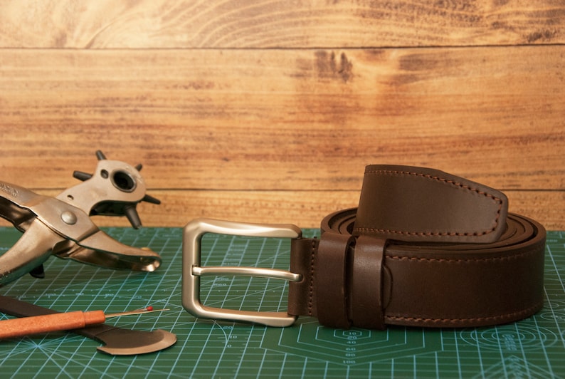 May include: A dark brown leather belt with a silver-toned metal buckle and matching stitching. Beside the belt lies a metal rotary leather hole punch tool and a small orange-handled seam ripper on a green cutting mat with grid markings.