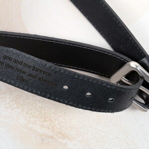 May include: A black leather belt featuring a silver-toned metal buckle and a personalised engraving on the inner side that reads: you and me forever, love you now and always, Ellen. The belt has visible stitching along the edges and holes.