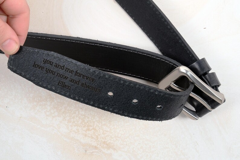 May include: A black leather belt featuring a silver-toned metal buckle and a personalised engraving on the inner side that reads: you and me forever, love you now and always, Ellen. The belt has visible stitching along the edges and holes.