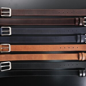 May include: A collection of eight leather belts arranged horizontally, featuring various colours including dark brown, navy blue, tan, and black. Each belt is equipped with a silver-toned metal buckle, a matching leather keeper, and five adjustment holes.