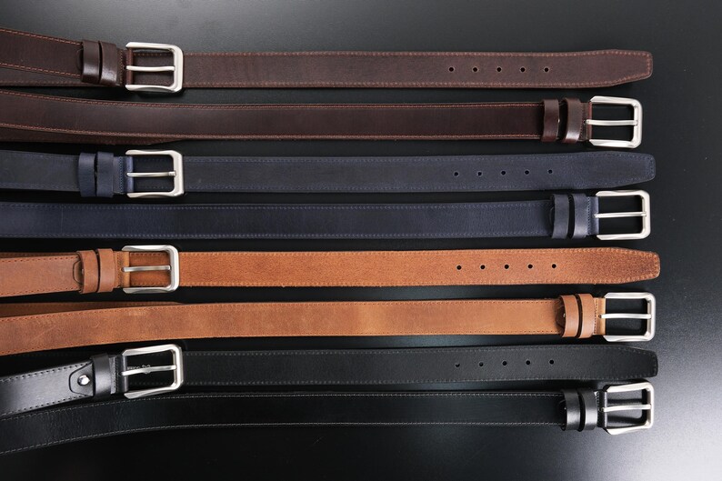 May include: A collection of eight leather belts arranged horizontally, featuring various colours including dark brown, navy blue, tan, and black. Each belt is equipped with a silver-toned metal buckle, a matching leather keeper, and five adjustment holes.
