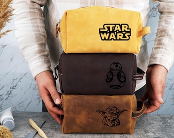 May include: Three stacked leather wash bags featuring different Star Wars characters. The top bag is yellow with the text "STAR WARS". The middle bag is brown with a black image of BB-8. The bottom bag is brown with a black image of Yoda.