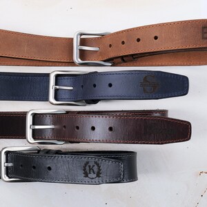 May include: Four leather belts with silver-toned metal buckles arranged horizontally. The top belt is tan with the letters BH, the second is navy blue with an S, the third is dark brown, and the bottom is black with a K inside a laurel wreath.