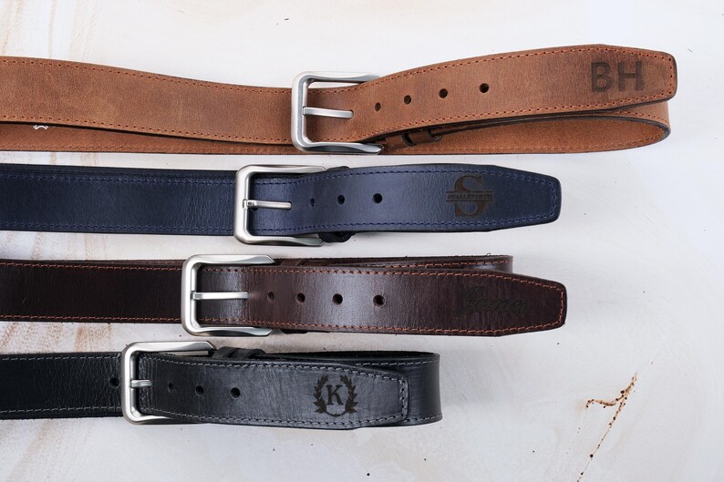 May include: Four leather belts with silver-toned metal buckles arranged horizontally. The top belt is tan with the letters BH, the second is navy blue with an S, the third is dark brown, and the bottom is black with a K inside a laurel wreath.
