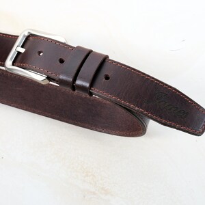 May include: A dark brown leather belt featuring a silver-toned metal buckle, two matching leather keepers, and visible stitching along the edges. The word James is embossed on the pointed end of the belt strap, which has a smooth finish.
