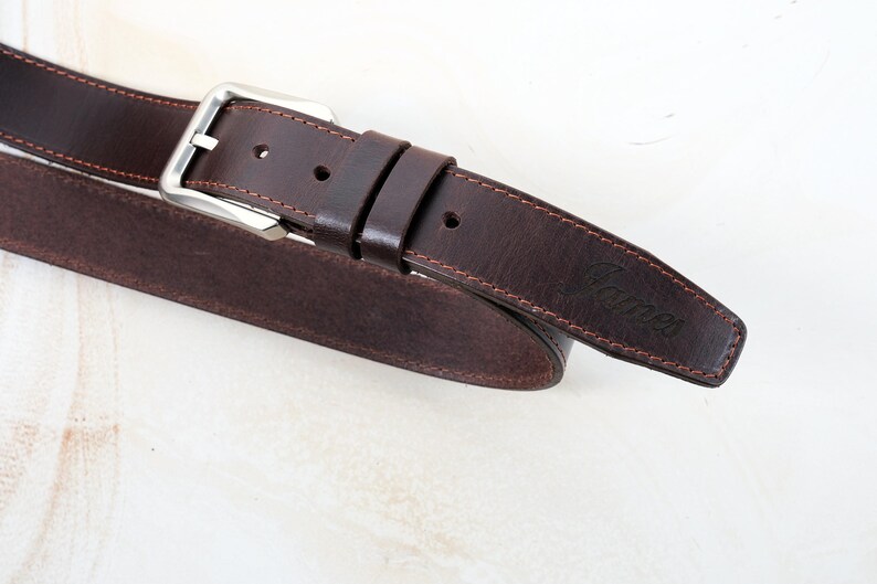 May include: A dark brown leather belt featuring a silver-toned metal buckle, two matching leather keepers, and visible stitching along the edges. The word James is embossed on the pointed end of the belt strap, which has a smooth finish.