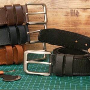 May include: A collection of leather belts in dark brown, black, and tan, each featuring a silver-toned metal buckle. A black leather belt is laid flat with a metal hole punch tool resting on it, alongside a seam ripper on a green cutting mat.