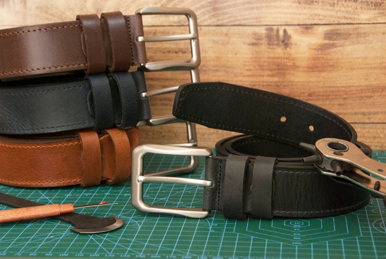 May include: A collection of leather belts in dark brown, black, and tan, each featuring a silver-toned metal buckle. A black leather belt is laid flat with a metal hole punch tool resting on it, alongside a seam ripper on a green cutting mat.