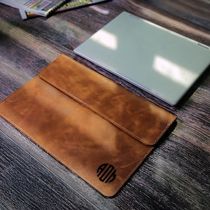 May include: A brown leather laptop sleeve with a monogrammed circle in the center. The sleeve is on a wooden surface with a silver laptop next to it.