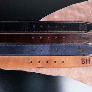 May include: Four leather belts in black, dark brown, navy blue, and tan, each featuring a row of adjustment holes and a personalised engraving on the end. The black belt shows a circular emblem, the brown belt says James, the blue belt has an S, and the tan belt displays the letters BH.