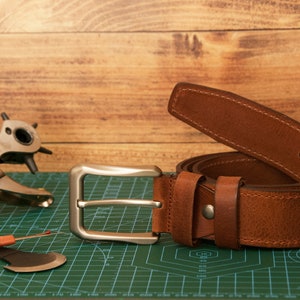 May include: A brown leather belt with a silver-toned metal buckle and matching leather loops. The belt features visible stitching along the edges. Beside the belt, a metal rotary hole punch tool and a small orange-handled seam ripper are visible.