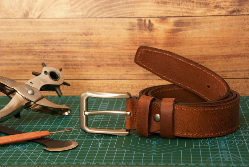 May include: A brown leather belt with a silver-toned metal buckle and matching leather loops. The belt features visible stitching along the edges. Beside the belt, a metal rotary hole punch tool and a small orange-handled seam ripper are visible.
