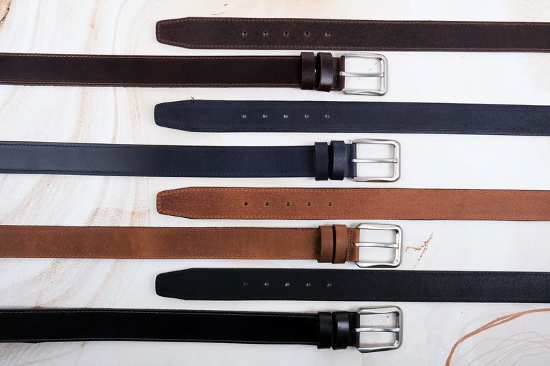 May include: A collection of four leather belts in dark brown, navy blue, tan, and black. Each belt features a smooth finish, a rectangular silver-toned metal buckle, two matching leather keepers, and a series of punched holes for adjustment.