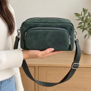 May include: A dark green rectangular crossbody bag with a smooth matte finish, featuring a front zippered pocket, a main zippered compartment, and a matching adjustable shoulder strap with silver-toned metal hardware and visible stitching.