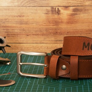 May include: A brown leather belt featuring a silver-toned metal buckle and the letters MG embossed on the strap. The belt is coiled and displayed alongside a metal leather hole punch tool and a small seam ripper on a green cutting mat.