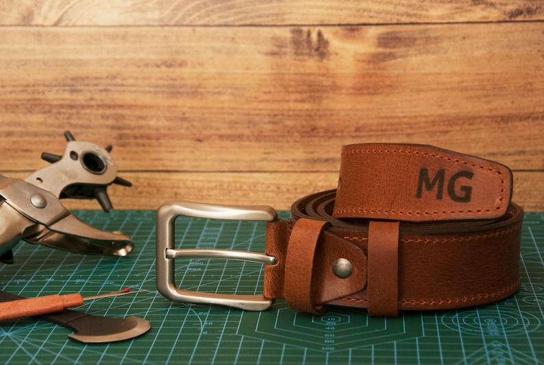 May include: A brown leather belt featuring a silver-toned metal buckle and the letters MG embossed on the strap. The belt is coiled and displayed alongside a metal leather hole punch tool and a small seam ripper on a green cutting mat.