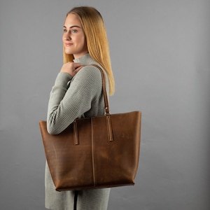 May include: A brown leather tote bag with a top zipper closure and two long handles. The bag is being held by a person wearing a gray sweater.