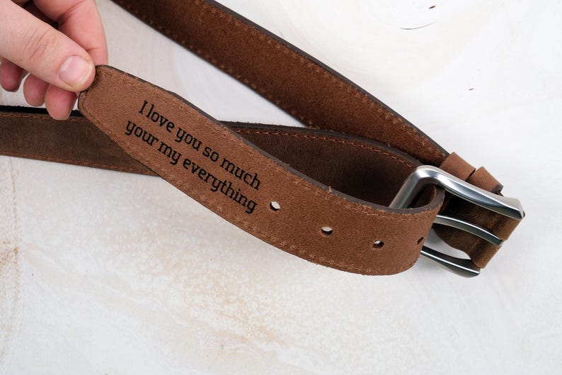 May include: A brown suede belt featuring a silver-toned metal buckle and a laser-engraved message on the inner strap that reads, I love you so much your my everything. The belt includes evenly spaced adjustment holes along the length of the strap.