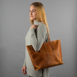 May include: A large, tan leather tote bag featuring a smooth, matte finish, a vertical center seam, and two long, thin shoulder straps attached with metallic rivets. The bag has a wide, open-top design with a structured, rectangular base.